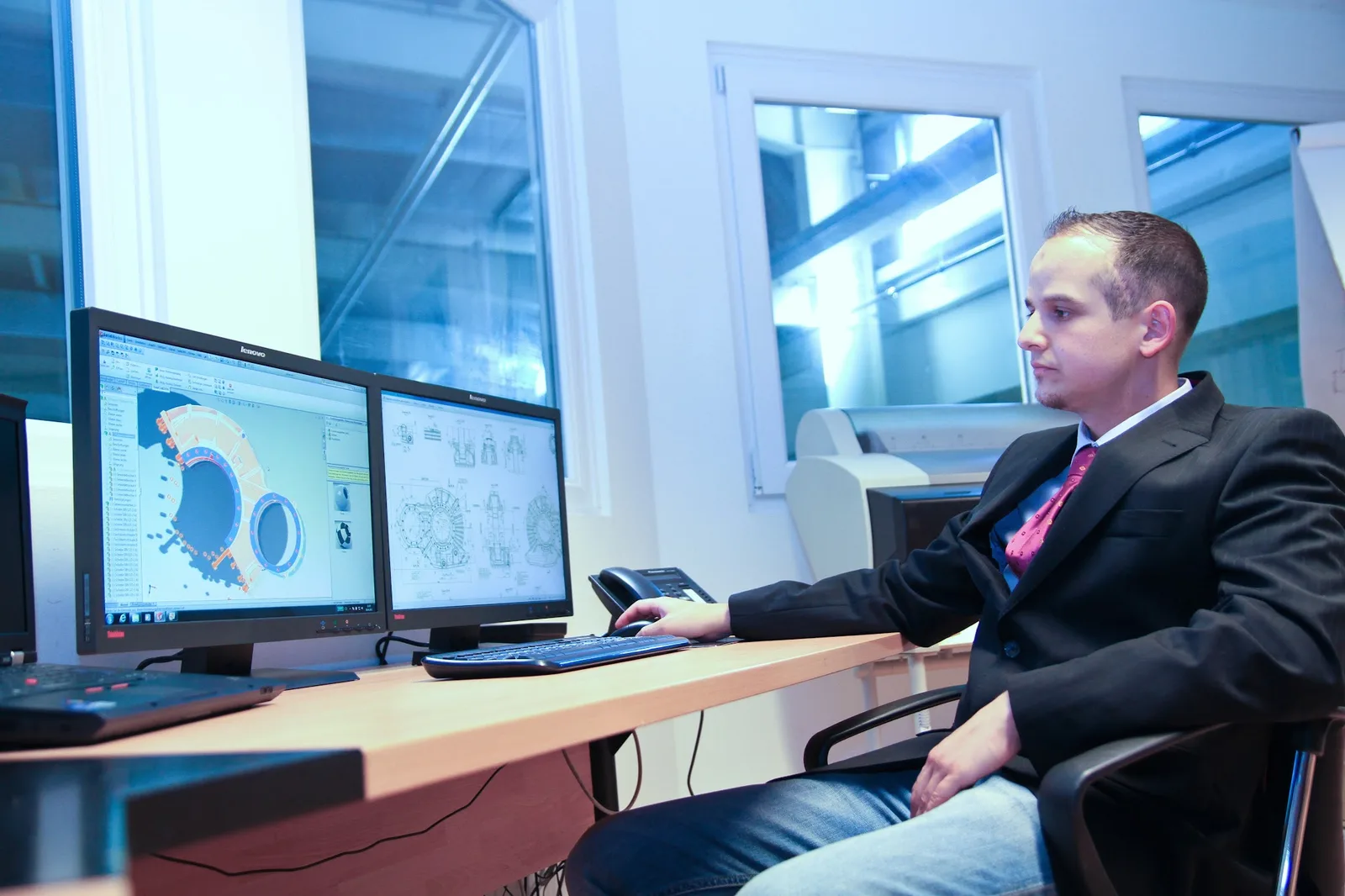 Mitarbeiter im Sakko am Arbeitsplatz mit zwei Bildschirmen, links ein 3D-Modell eines Gehäuses, rechts eine technische Zeichnung