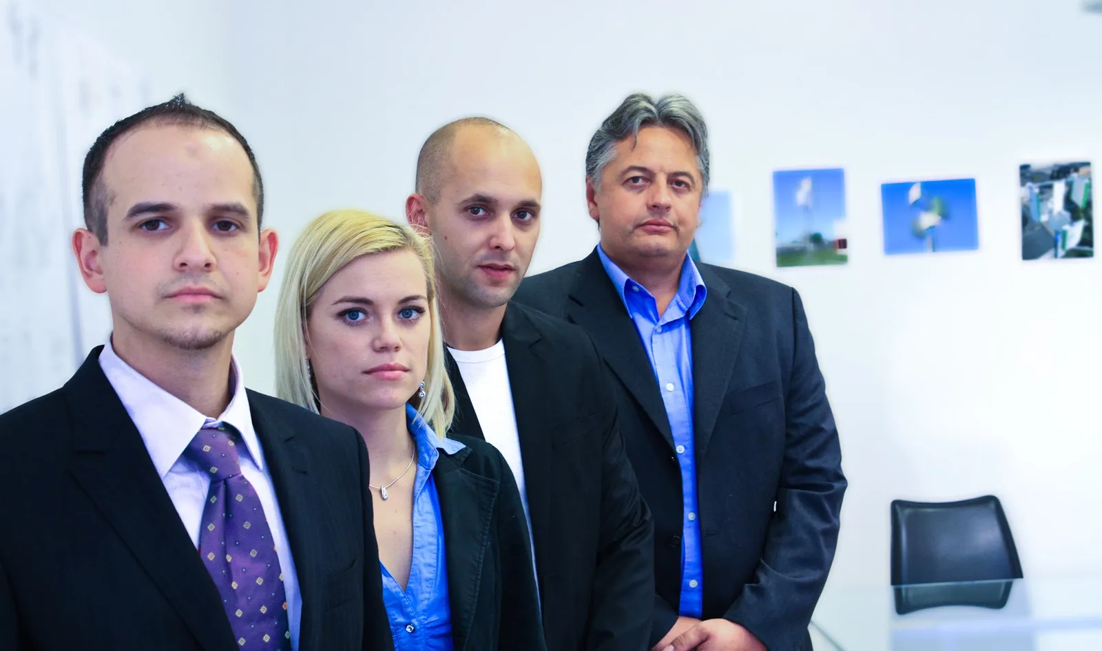 Vier Personen im Sakko, nebeneinander vor einer weißen Wand mit Fotos