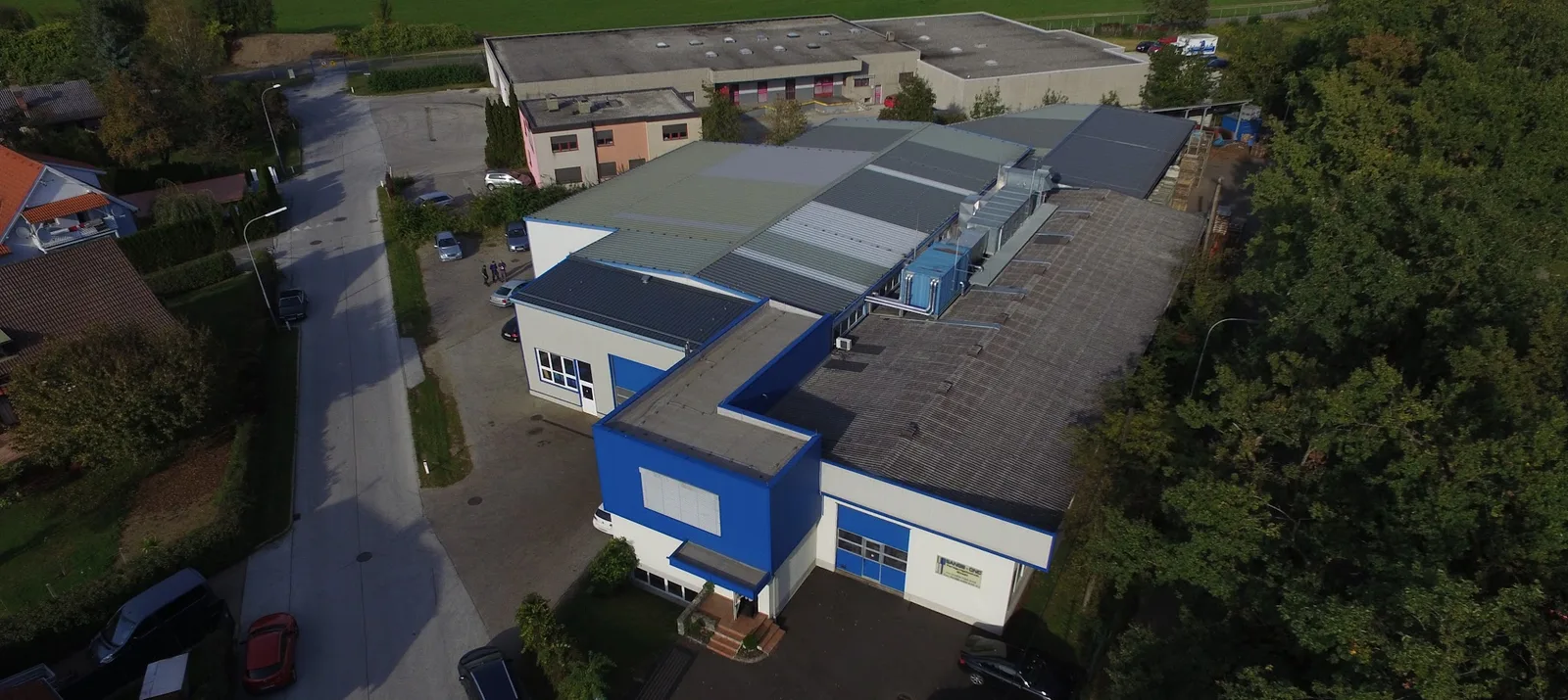 Luftbild des Firmengebäudes mit blau-weißer Fassade, Hallendächern und Parkplatz im Gewerbegebiet